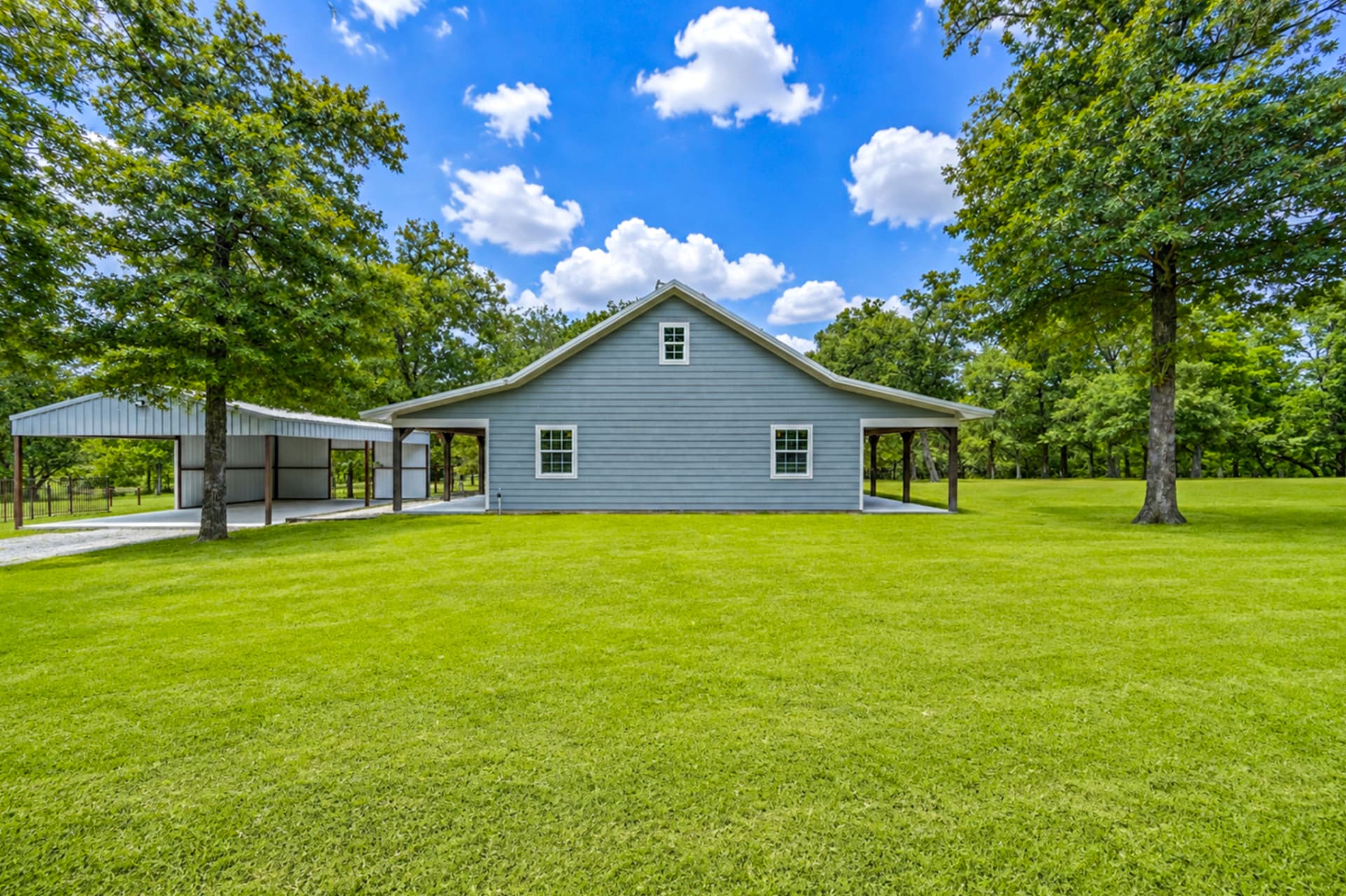 Front of the blue farmhouse with attached carport and pole barn under mature oaks